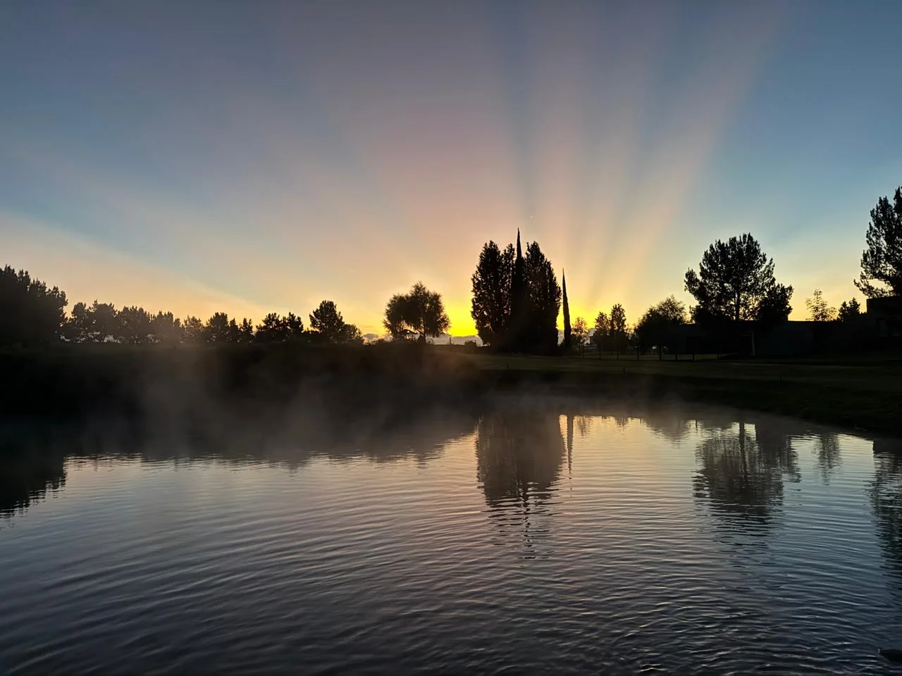 Lago al amanecer con reflejos, bruma y árboles.