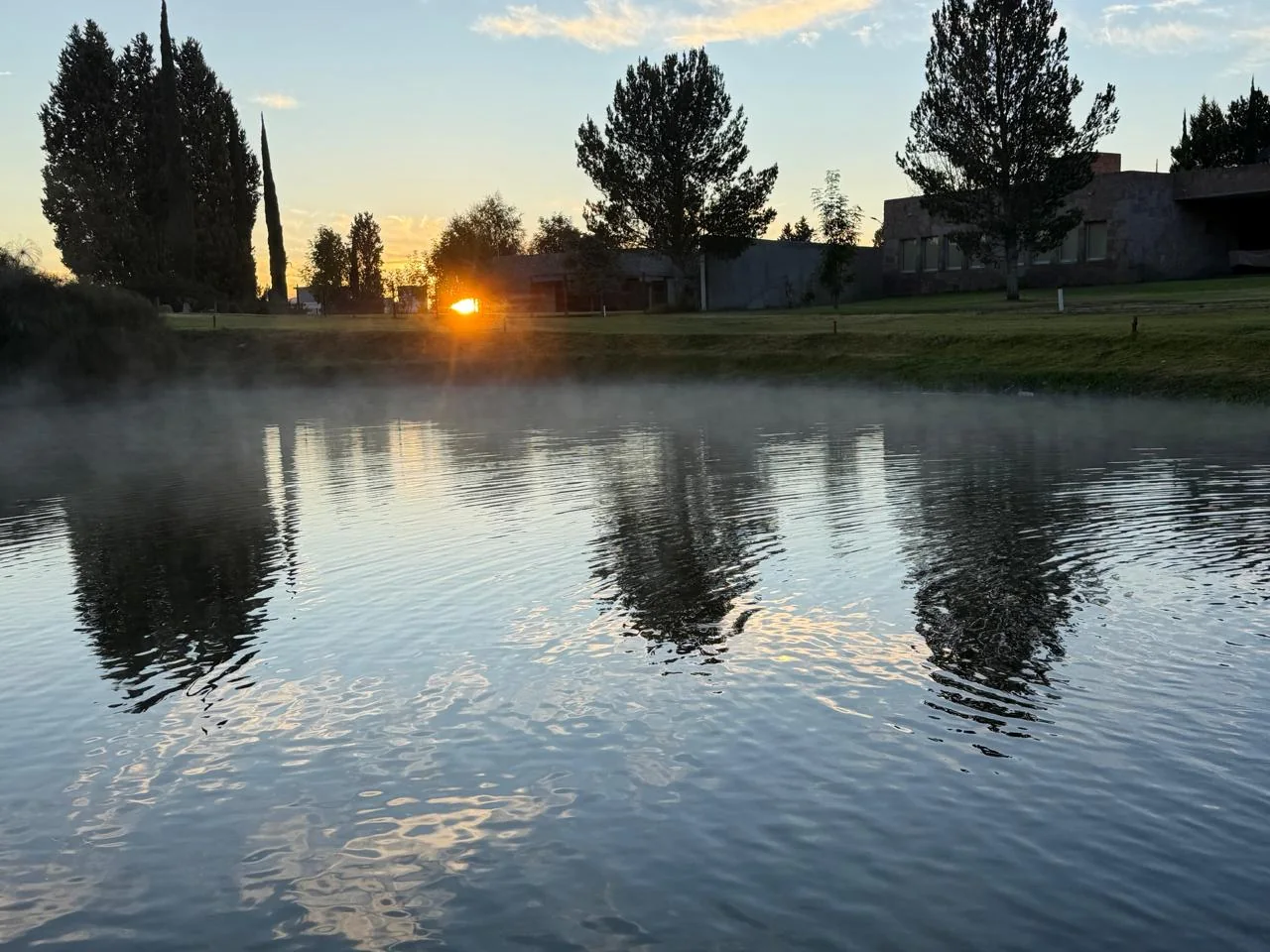 Lago al amanecer con reflejos, bruma y árboles.